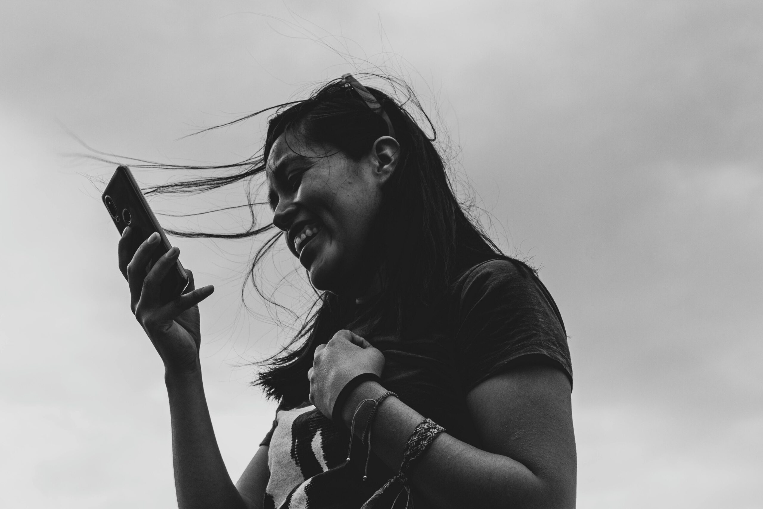 Black and white portrait of a smiling woman using a smartphone outdoors, exuding happiness and modern lifestyle.