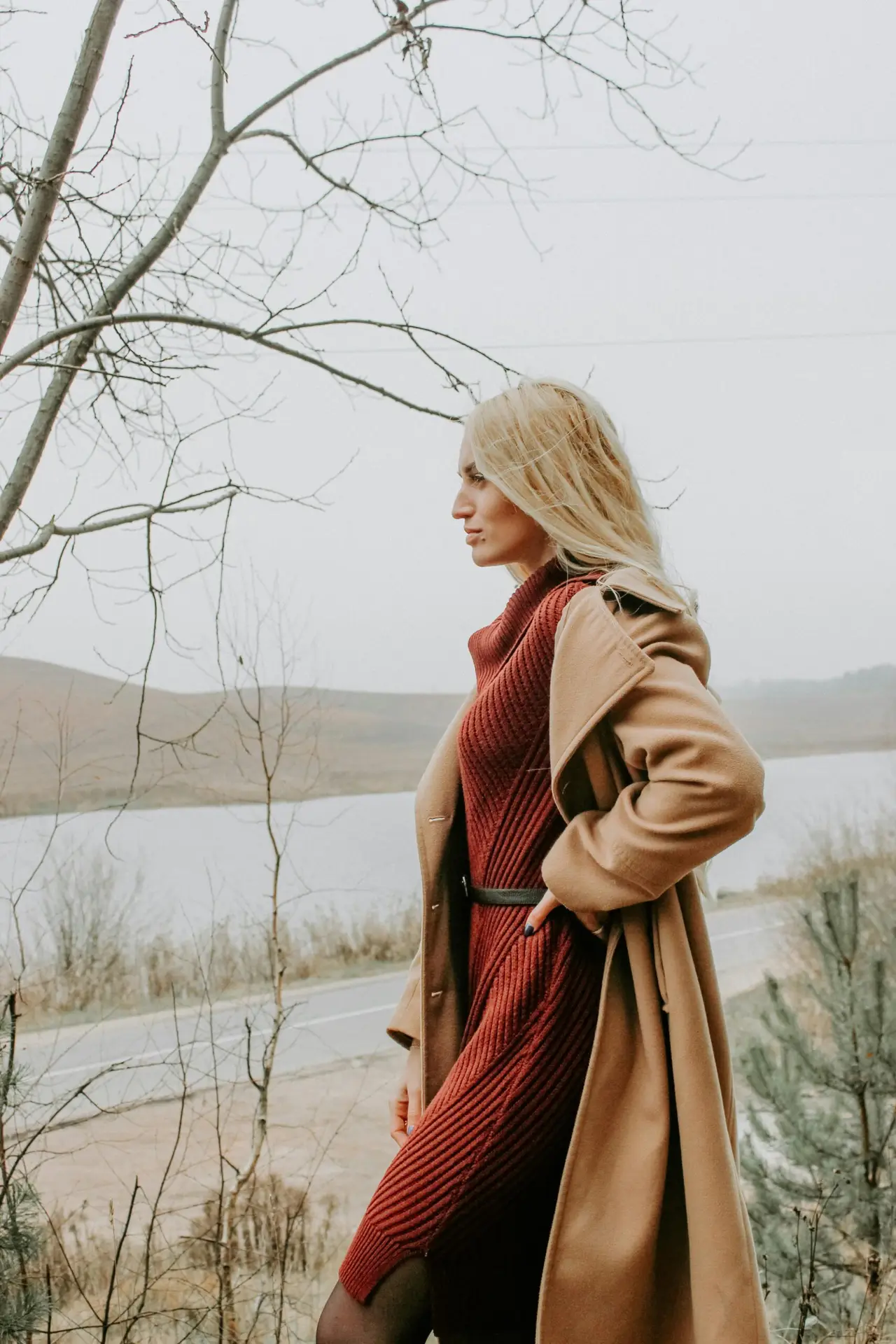 Elegant woman in a brown coat standing by a tranquil lake during autumn.